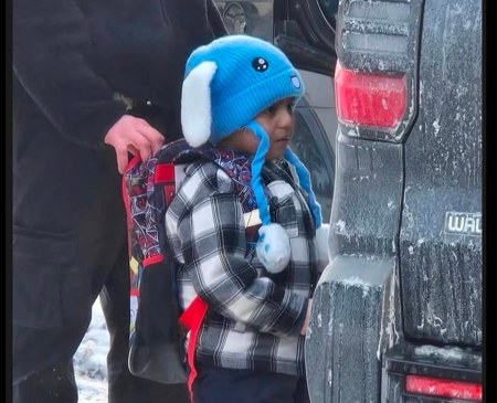 Liam, a five-year-old boy in a blue bunny hat and plaid coat, standing beside a vehicle in the snow